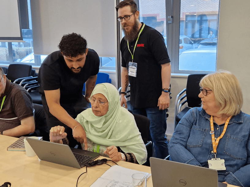 A training session in progress, with an older tenant using a laptop being aided by two trainers, whilst a second tenant looks on