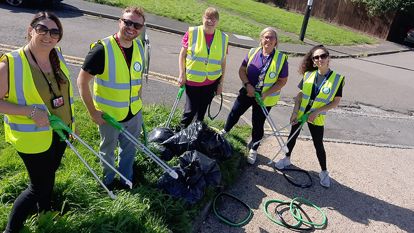 A team of litter pickers wearing high-vis vests and holding litter pickers, with hoops and filled rubbish sacks, standing on a grass verge