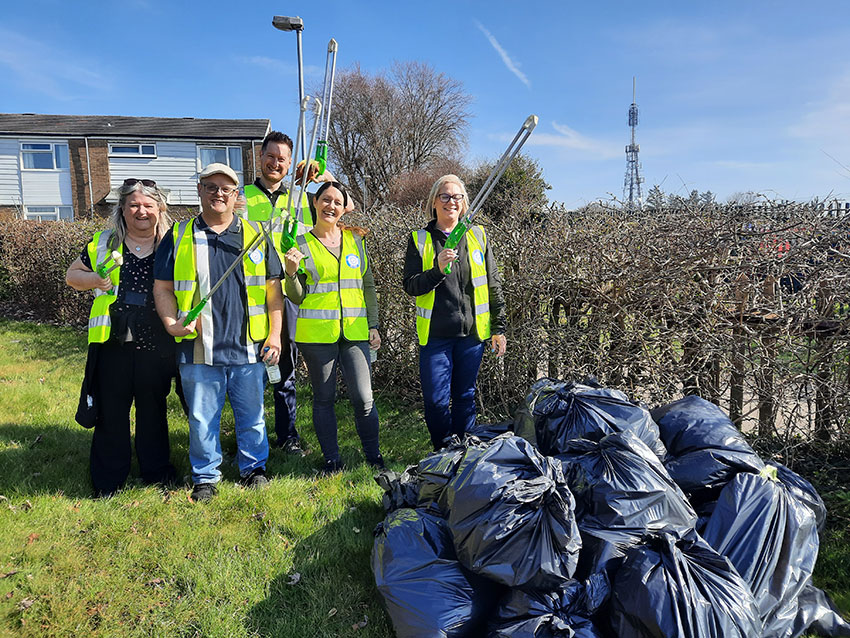 A team of litter pickers wearing high-vis vests and holding litter pickers, with filled rubbish sacks, standing on a grass verge