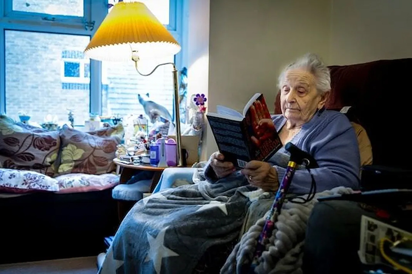 An older woman sat reading by the light of a standard lamp